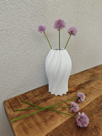 White vase with purple flowers on a wooden surface against a plain wall