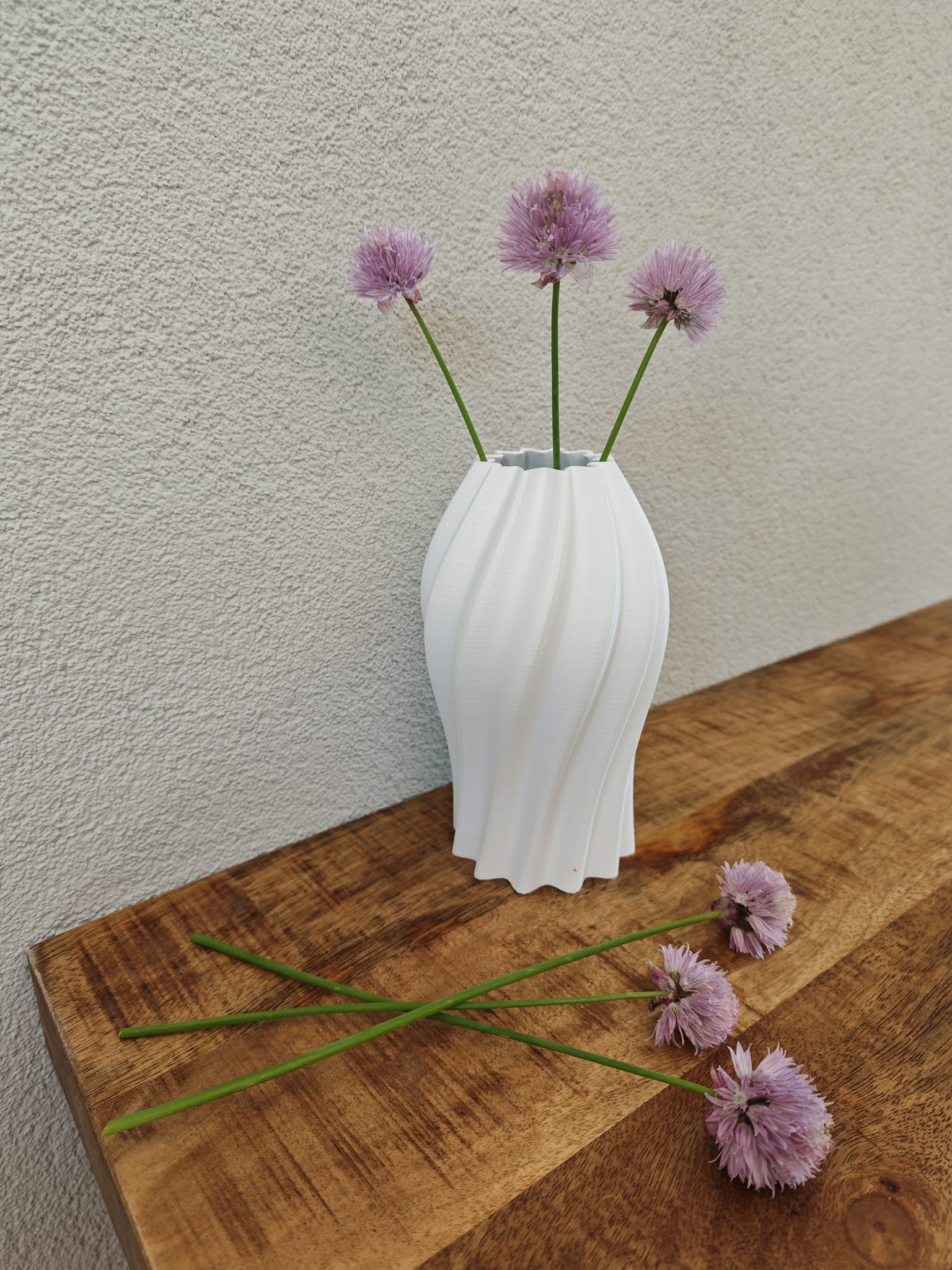 White vase with purple flowers on a wooden surface against a plain wall